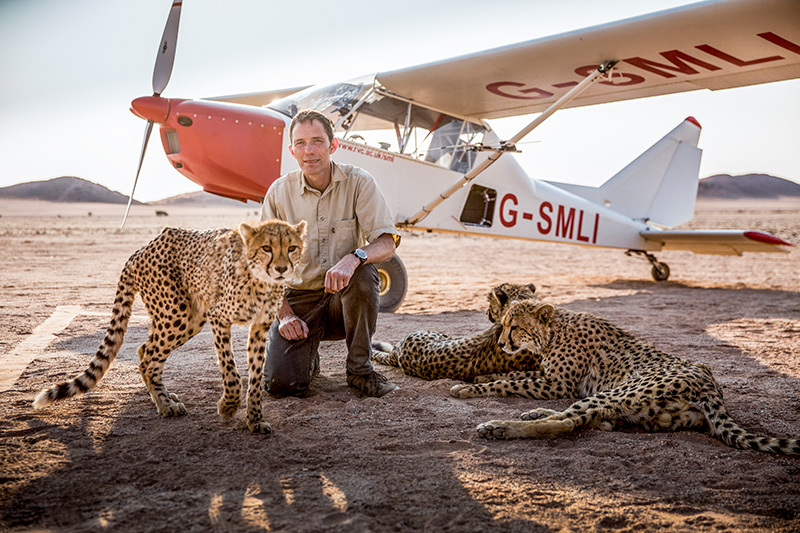 Alan Wilson with three cheetahs in front of his light aeroplane in a desert landscape